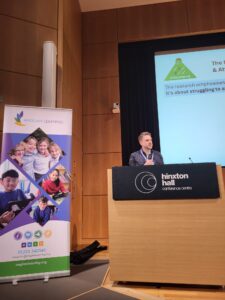 A man in a navy shirt is standing behind a podium on a stage. Next to him is a large roller banner which has images of pupils and Anglian Learning's logo. Behind the man is a large screen which has a PowerPoint presentation on the screen.