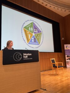 A lady stands behind a podium on a stage. Behind her is a large screen which shows a presentation slide with a colourful image.