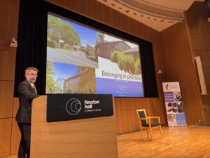 A man in a grey suit is standing behind a podium on a stage. Behind him is a large screen which has a PowerPoint Presentation slide of images of a school.