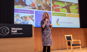 A lady in a blue dress is standing on a stage holding a microphone to her mouth. Behind her is a large screen which shows a PowerPoint presentation.