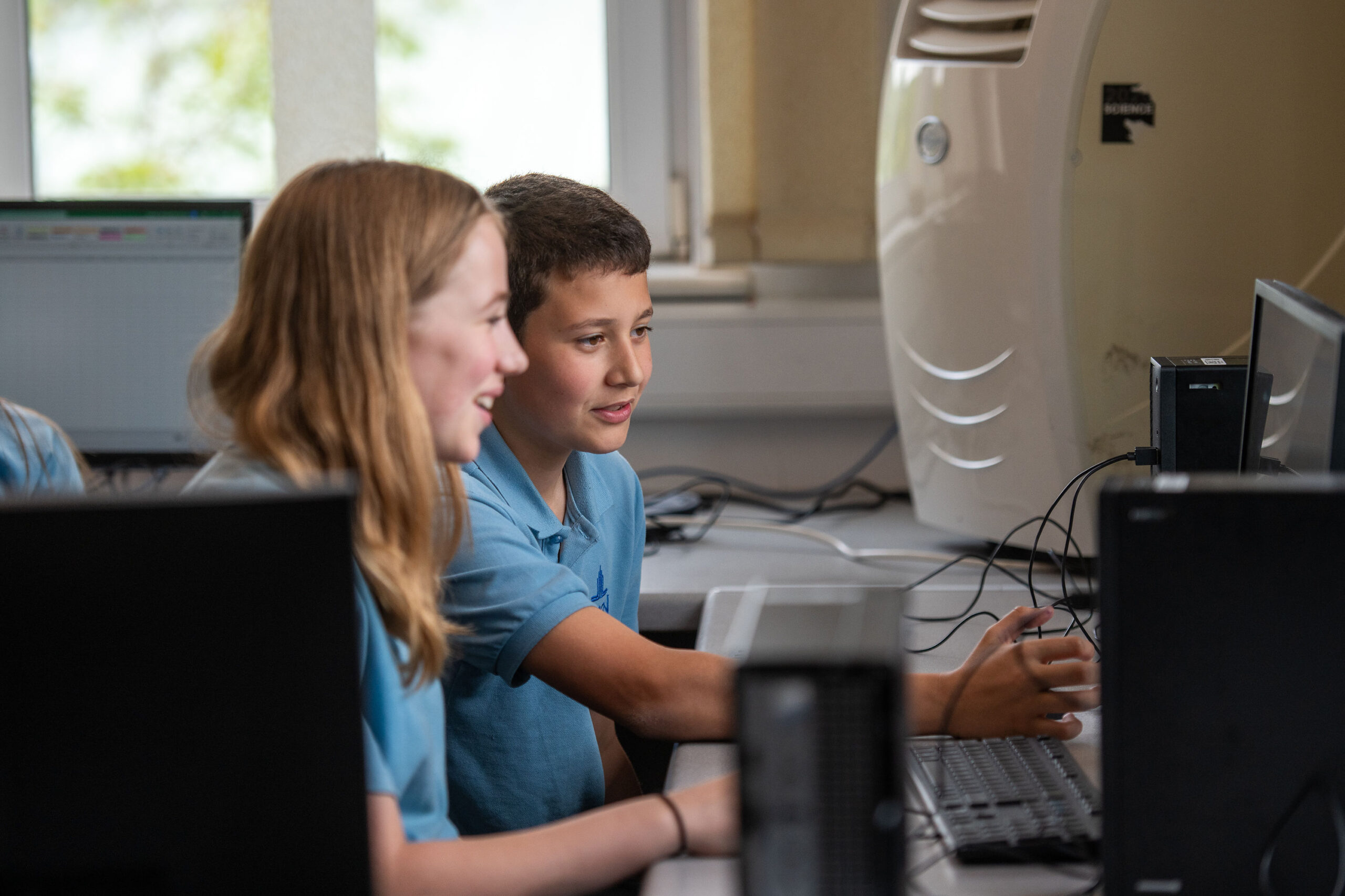 Two pupils are sitting at a computer. One girl is holding the mouse and staring at the screen. One boy is pointing to the computer screen. Both are wearing blue polos with the school's logo embroidered on the top.