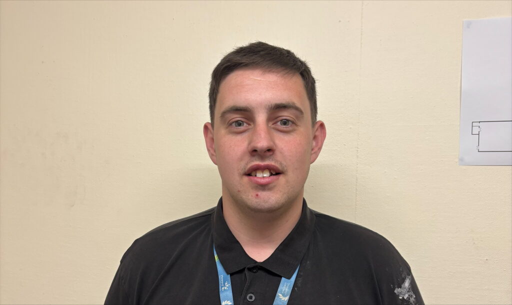 A man is standing in front of the camera smiling. He is wearing a blue polo neck and blue lanyard.