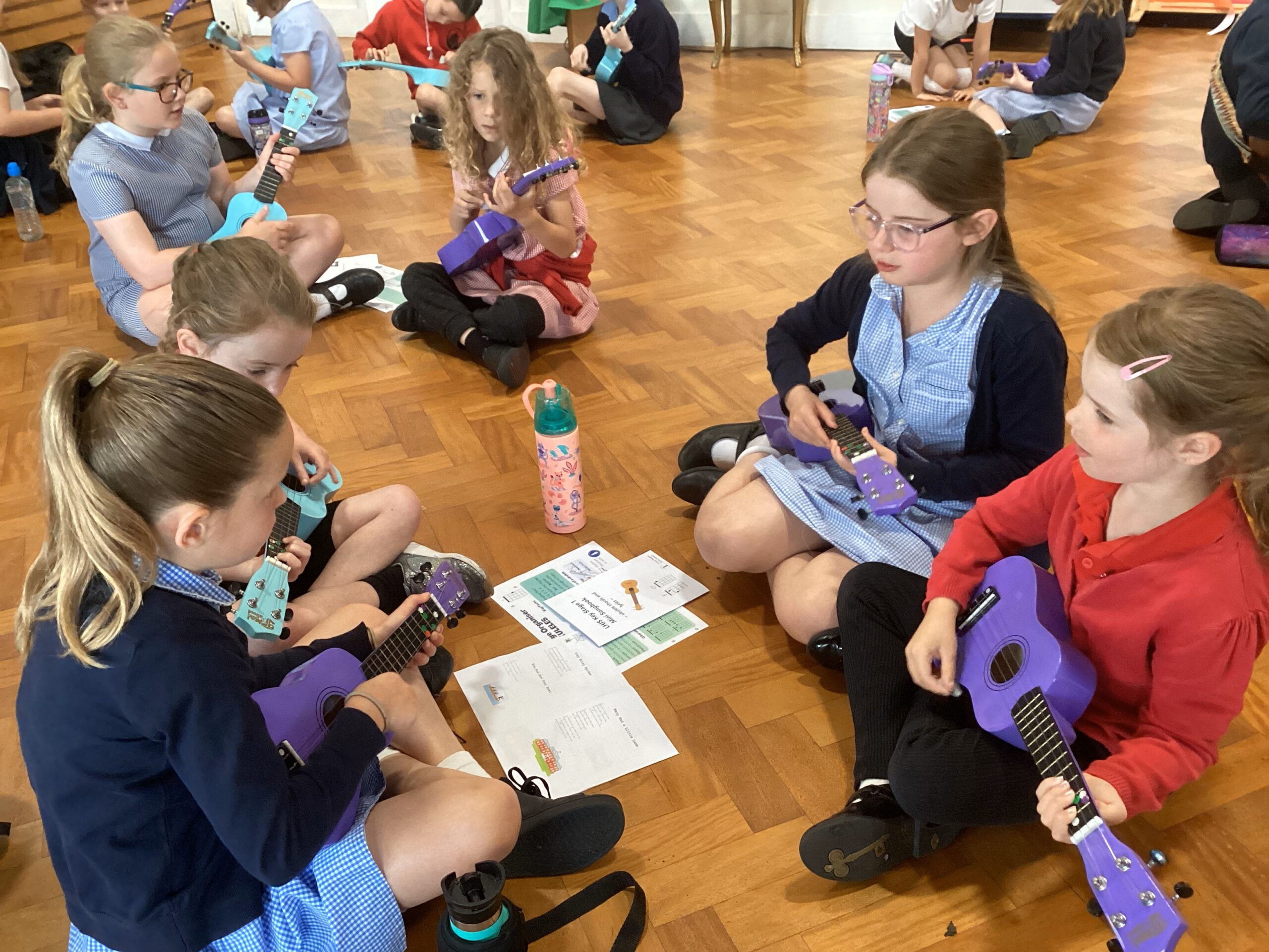 Six children are sitting on the floor facing each other. They are each holding a purple ukulele instrument. They are facing each other whilst they practice playing the instrument.