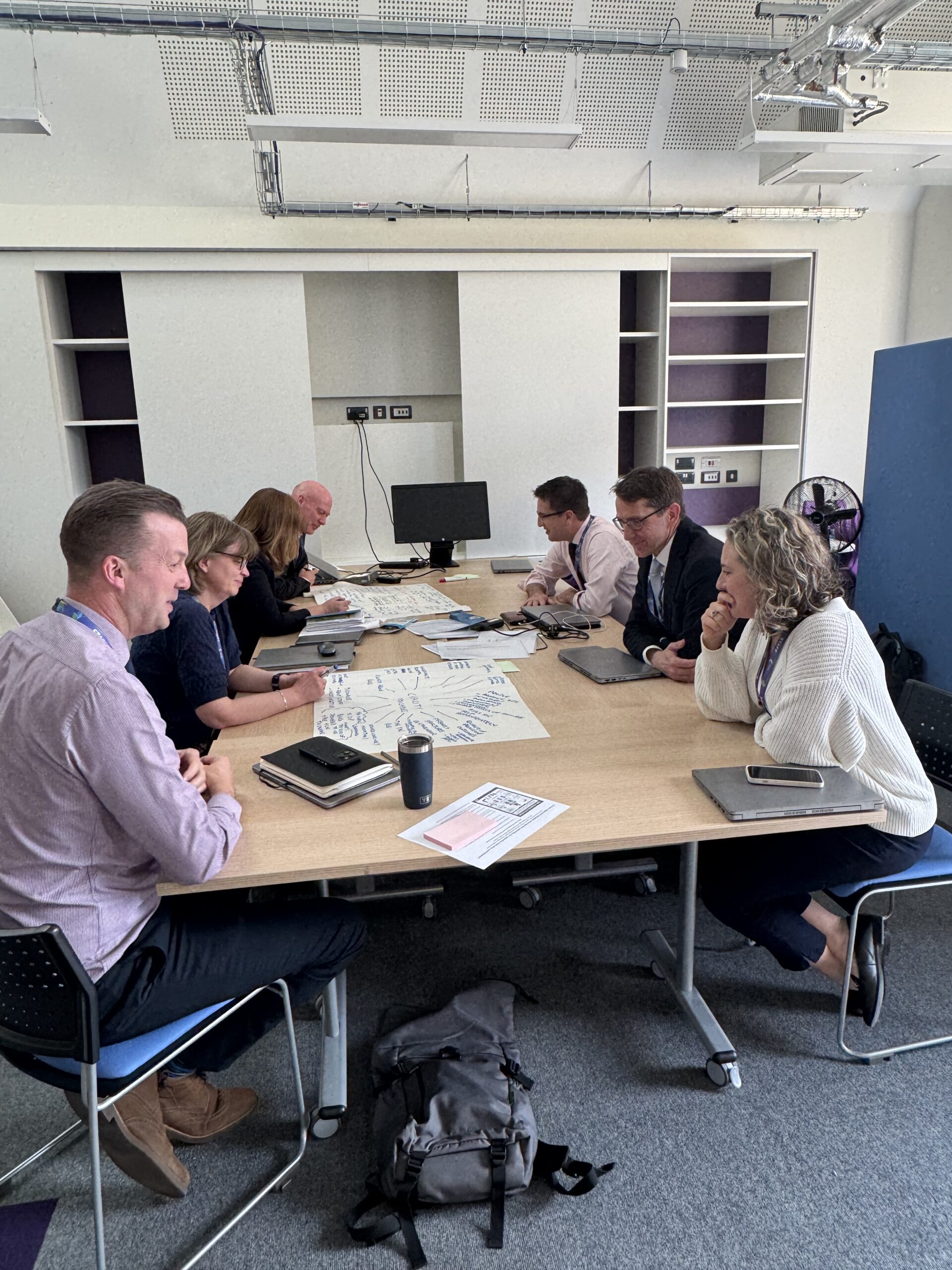 A large group of people are sitting at a desk facing each other. They are having a discussion with each other.