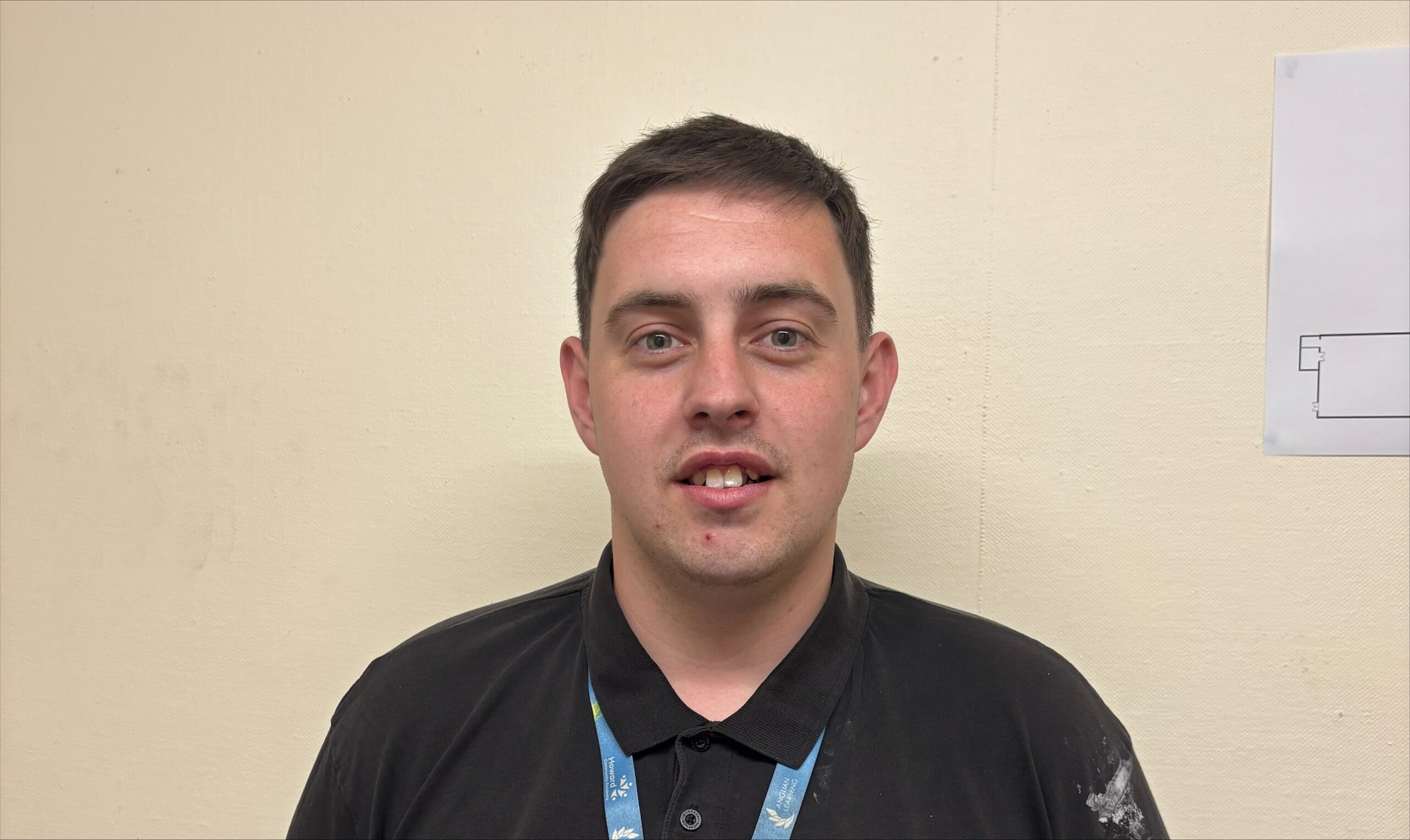A man is standing in front of the camera smiling. He is wearing a blue polo neck and blue lanyard.